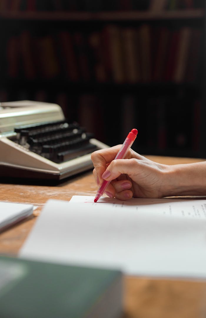 A close-up shot of a hand writing on papers with a red pen beside a vintage typewriter.